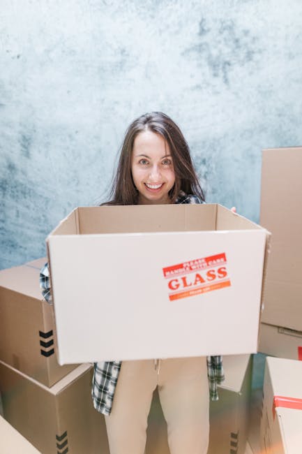A woman with long brown hair, smiling and looking directly at the camera, is standing inside a room with a light grey textured wall. She is holding an open cardboard box without a lid, which has a red